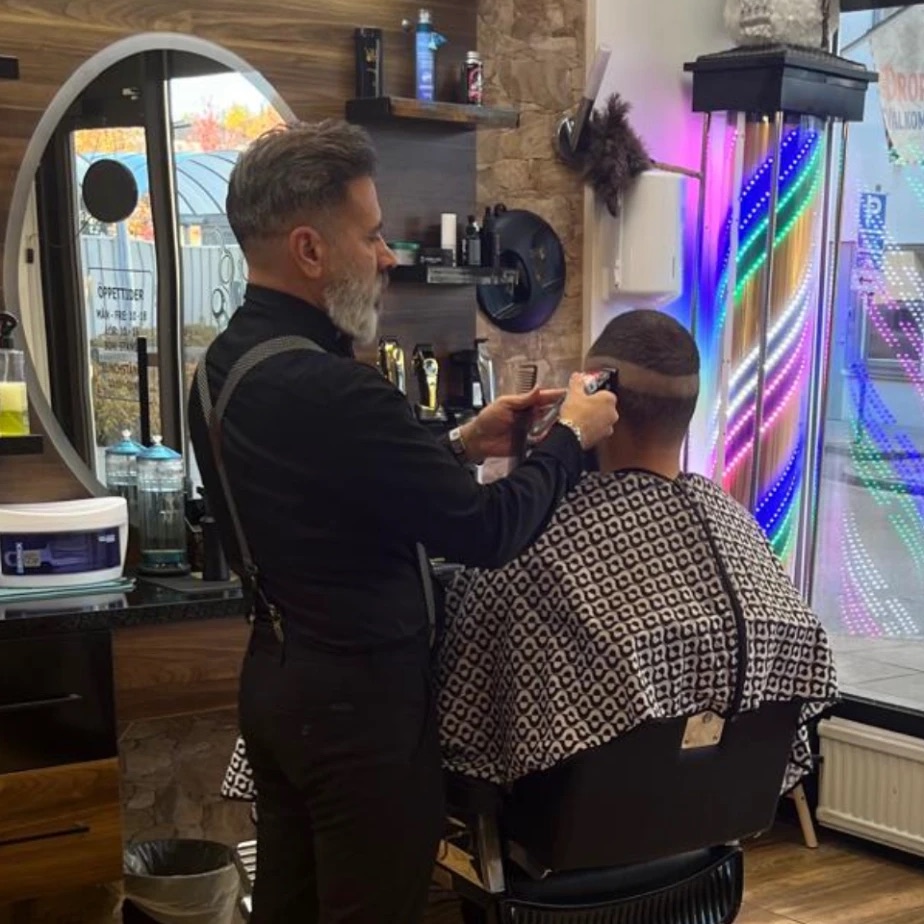Frisören klipper en fade med maskin och kam, med salongens upplysta barberarstolpe i bakgrunden